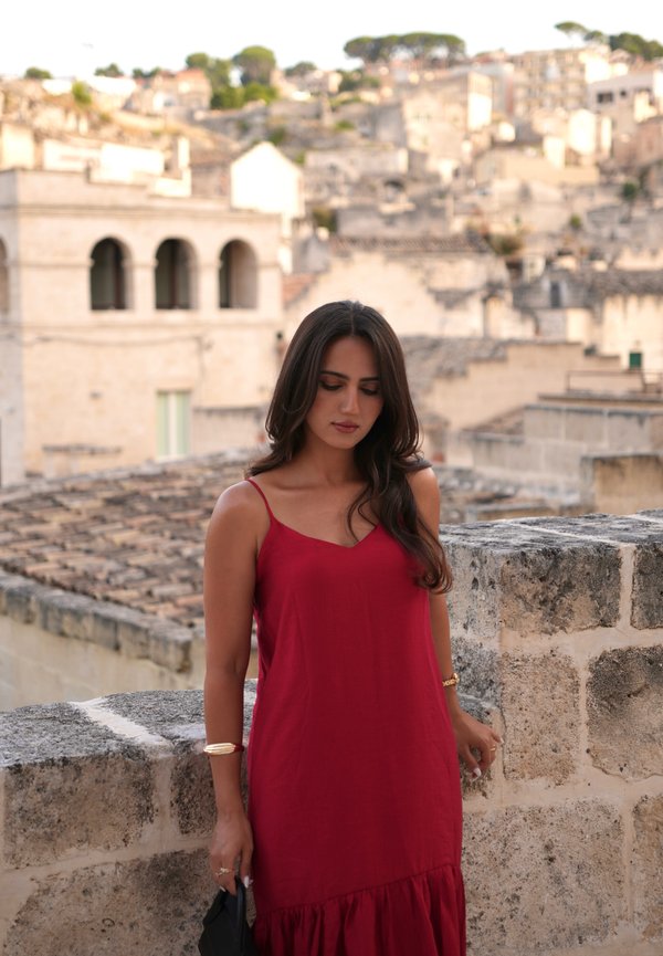 Frau in rotem Kleid steht an einer Steinmauer mit historischen Gebäuden und einem Hügel im unscharfen Hintergrund.