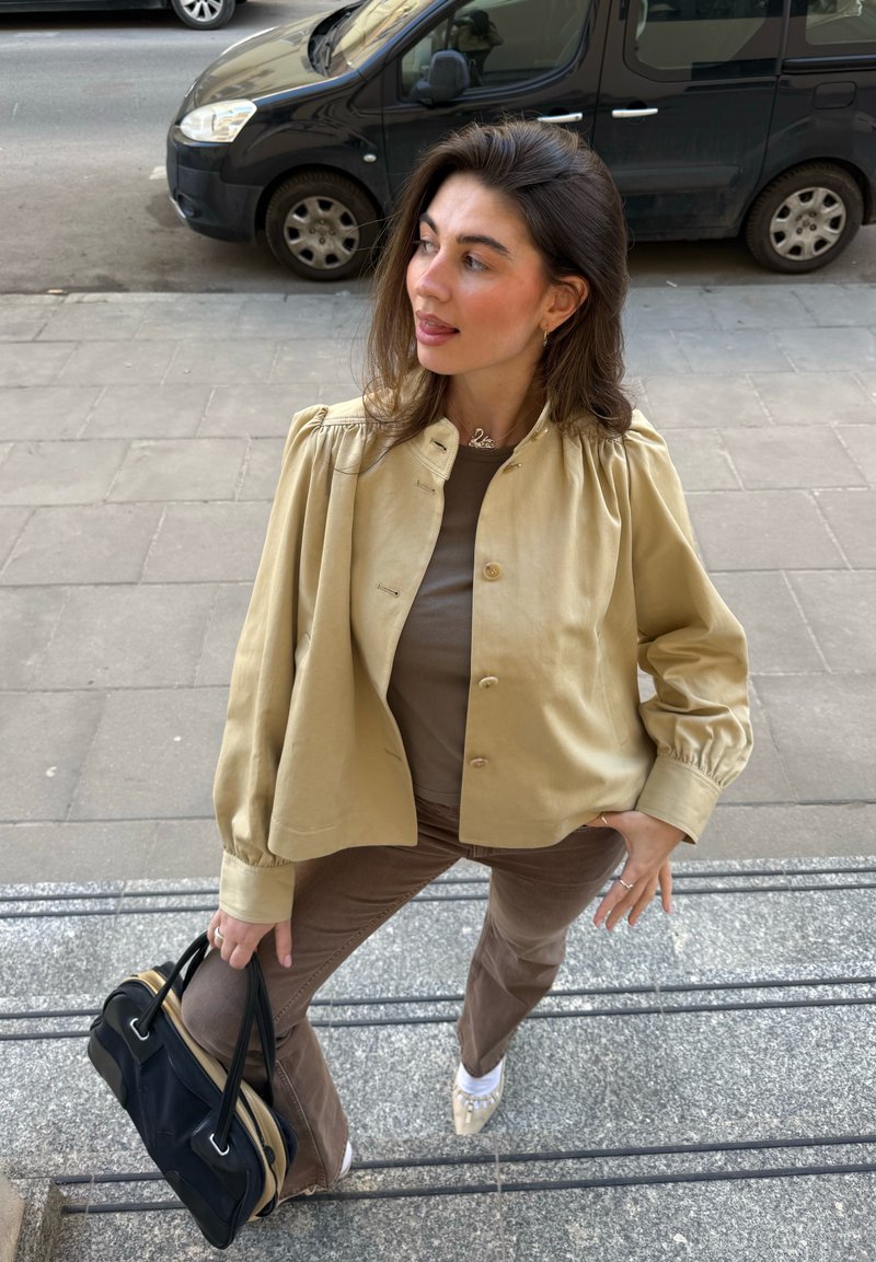 Γυναίκα με καστανά μαλλιά, που φοράει μια μπεζ μπουφάν και καφέ παντελόνι, κρατά μια μαύρη τσάντα, στέκεται σε εξωτερικά σκαλοπάτια κοντά σε παρκαρισμένα αυτοκίνητα.