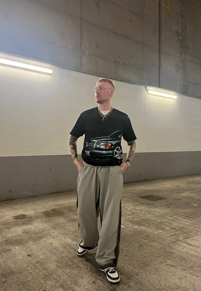 Camiseta negra con un gráfico de un coche; pantalones de chándal grises sueltos; zapatillas blancas y negras; cabello corto; gafas; ambiente interior con luces en el techo.