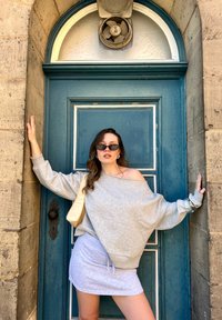 Sweatshirt cinza à mostra os ombros combinado com uma saia mini cinza clara. Os acessórios incluem óculos de sol pretos pequenos e uma mala de ombro bege. Fundo de porta azul.