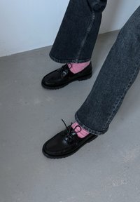 Chaussures en cuir noir à lacets, surface texturée et semelles épaisses, port ées avec des chaussettes roses. Portées avec un jean gris foncé à jambes larges.