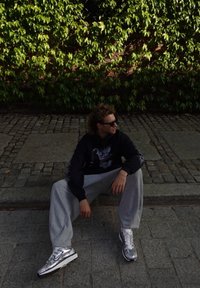 Person sitting on a cobblestone path, wearing a black hoodie with a graphic design, gray sweatpants, and silver sneakers. Green foliage in background.