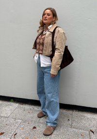 Woman with beige jacket over brown argyle vest and white shirt, blue jeans, brown shoes, and a dark brown shoulder bag against a gray wall.