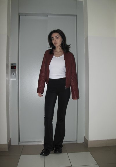 Mujer de pie frente a una puerta cerrada de ascensor, vestida con una chaqueta roja, camiseta blanca, pantalones negros y zapatos negros en el interior.