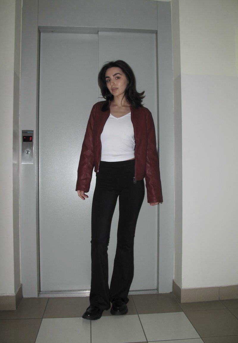 Ženska stoji pred zaprtimi vrati dvigala, oblečena v rdečo jakno, belo majico, črne hlače in črne čevlje v notranjosti.