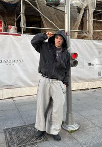 Hoodie noir court, sweatpants gris surdimensionnés avec des détails de poches, chaînes en argent superposées et bottes noires ; debout près d'un feu de circulation.