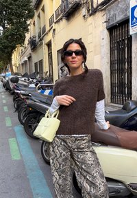 Femme portant des lunettes de soleil foncées, un pull marron à manches courtes, un haut blanc à manches longues, un pantalon à imprimé serpent, tenant un sac à main jaune clair dans une rue de la ville.