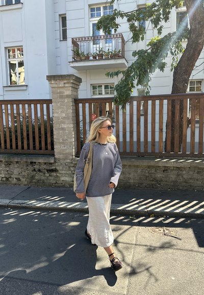 Camisa de rayas de manga larga en azul marino y blanco, falda de lino blanca, sandalias negras, llevando un bolso beige, caminando por una acera soleada.