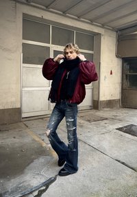 Red bomber jacket, black scarf, distressed blue flared jeans, and black platform shoes. Model poses outdoors beside a textured surface.