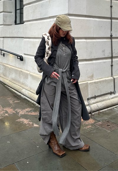 Persona con gorra verde, abrigo largo oscuro, blusa gris con cinturón, pantalones anchos grises y botas marrones de punta cuadrada, de pie junto a la esquina de un edificio de piedra.