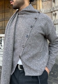 Homme portant un pull gris texturé boutonné sur un pantalon à carreaux avec une boucle de ceinture argentée détaillée, debout à l'extérieur près d'un mur en pierre orné.