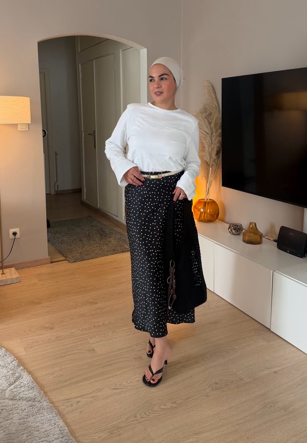 Mujer con una blusa blanca de manga larga, falda negra con lunares, pañuelo beige en la cabeza y tacones negros, de pie en un salón moderno.