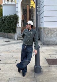 Casaco verde com fecho éclair e acessórios em prateado, calças largas azuis escuras, e um boné bege. A pessoa está de pé casualmente ao lado de um poste, segurando um copo.