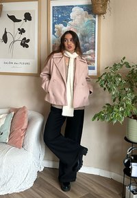 Hellrosa, doppelt knöpfbarer Wollmantel über einem beigen Pullover, kombiniert mit schwarzen, weit geschnittenen Hosen und schwarzen Knöchelstiefeln. Weiße Strickschal.