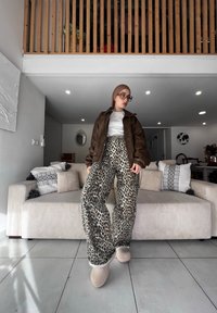 Hnedá kožená bunda, biela tričko a nohavice so širokými nohavicami v leopardom vzore. Mäkké béžové topánky. Pohodlná pohovka s textúrovanými vankúšmi v modernom interiéri.