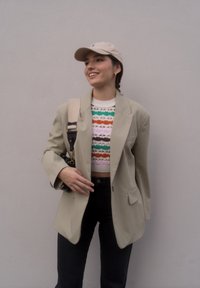 Blazer oversized em bege claro, suéter tricotado cropped multicolor, jeans escuros, boné bege e mala de ombro com alça. Conjunto casual em camadas.