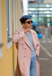 Trench coat roz pal cu butoane aurii, tricou cu dungi albastre, beret albastru și ochelari de soare închiși; stând în fața unei pereți galbene.