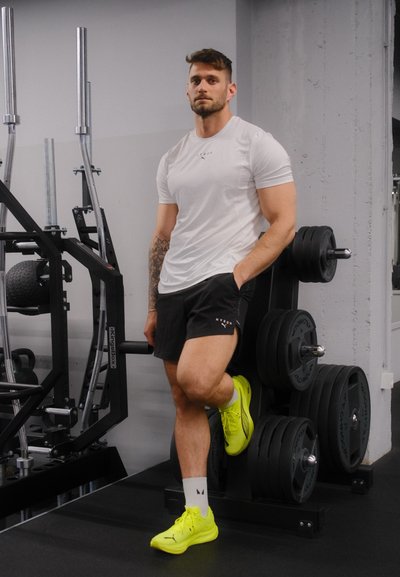 Camiseta atlética blanca, pantalones cortos negros y zapatillas brillantes amarillas. De pie junto a equipo de levantamiento de pesas en un gimnasio.