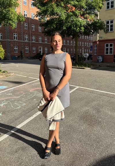 Vestido sin mangas gris con una silueta ajustada, sosteniendo un suéter de tono claro. Zapatos negros con correas, de pie sobre una superficie pavimentada.