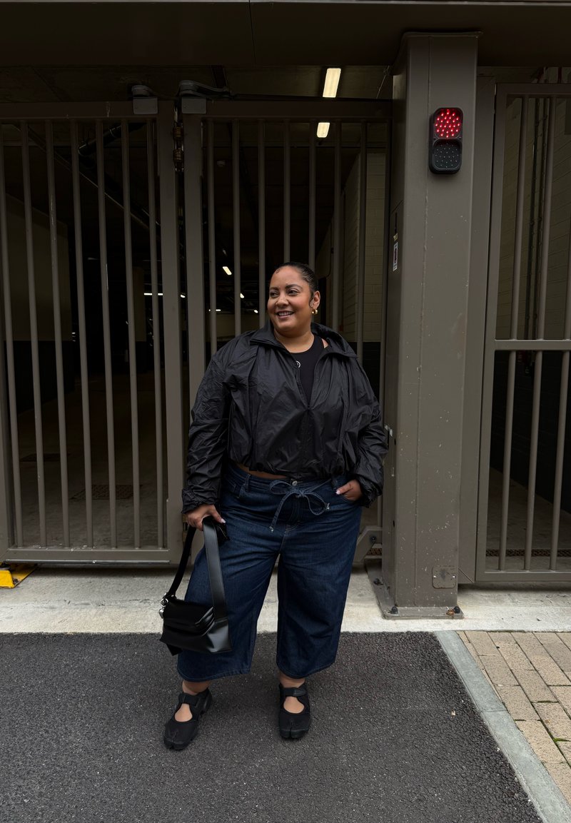 Black cropped jacket, black top, wide-leg dark denim jeans with a drawstring waist, and black slip-on shoes. Carrying a small black bag.
