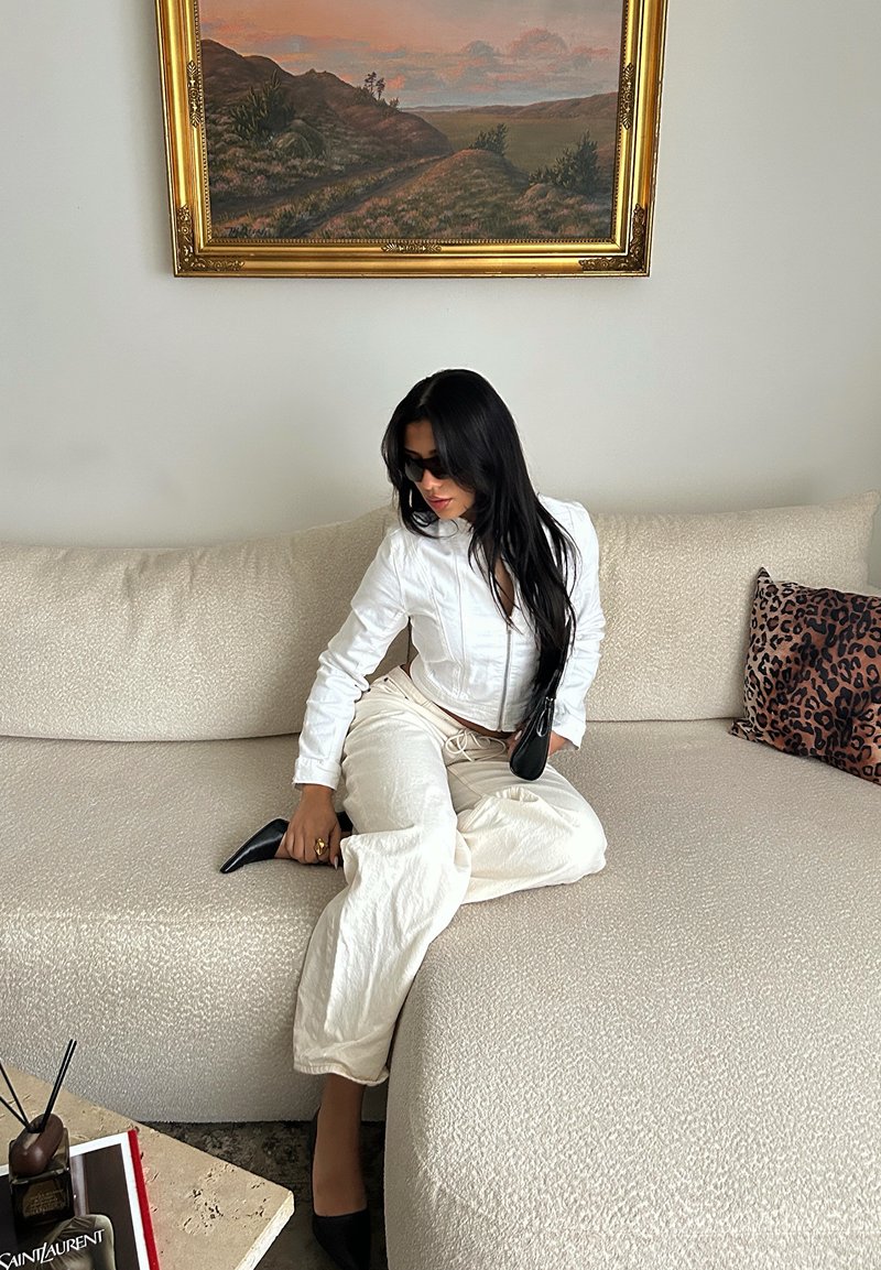 Camicia bianca corta, pantaloni crema a gamba larga, scarpe nere con tacco. Seduta su un divano chiaro dalla trama evidente, con un cuscino leopardato, dietro un quadro paesaggistico incorniciato.