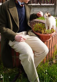 Zelený vlnený kabát na námorníckom svetri, krémové nohavice, sediaci na tehlovej stene s bielou mačkou pri kvetináči. V pozadí trávnik.