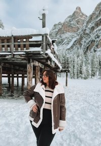 Braune Lederjacke mit weißem Schaffellkragen, über einem beigen gestreiften Pullover getragen, vor einer schneebedeckten Landschaft mit einem Holzgebäude.
