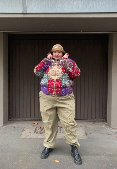 Chaqueta acolchada de patchwork colorido con varios patrones, cierre con cremallera y capucha, combinada con pantalones khaki holgados y botas negras.