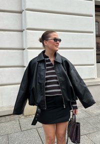 Czarna skórzana kurtka bomberka z szerokim kołnierzem, noszona na paskowanym crop topie i czarnej mini spódniczce, trzymająca plecioną ciemną torebkę.