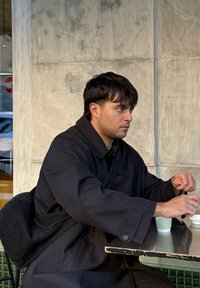 Νεαρός άνδρας με σκούρο παλτό ανακατεύει ένα ποτό ενώ κάθεται σε τραπέζι εξωτερικού καφέ μπροστά σε υφή τσιμεντένιου τοίχου.