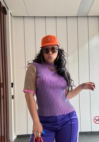 Fialový a růžový pruhovaný pletený top s krátkými, barevnými rukávy; oranžová čepice; černé oversized sluneční brýle; fialové kalhoty; kabelka v ruce.