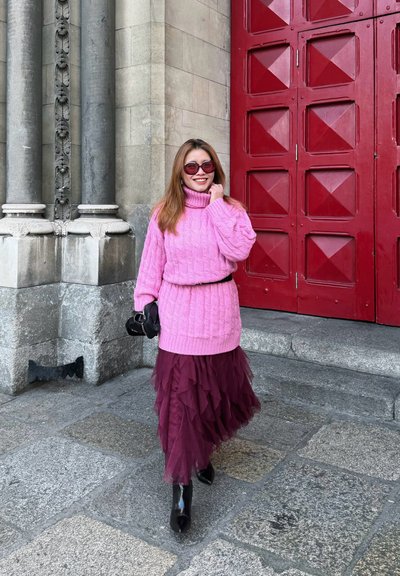Mujer sonriente, vestida con suéter rosa y falda de tul burdeos, caminando sobre un pavimento de piedra cerca de columnas ornamentadas y una gran puerta roja.