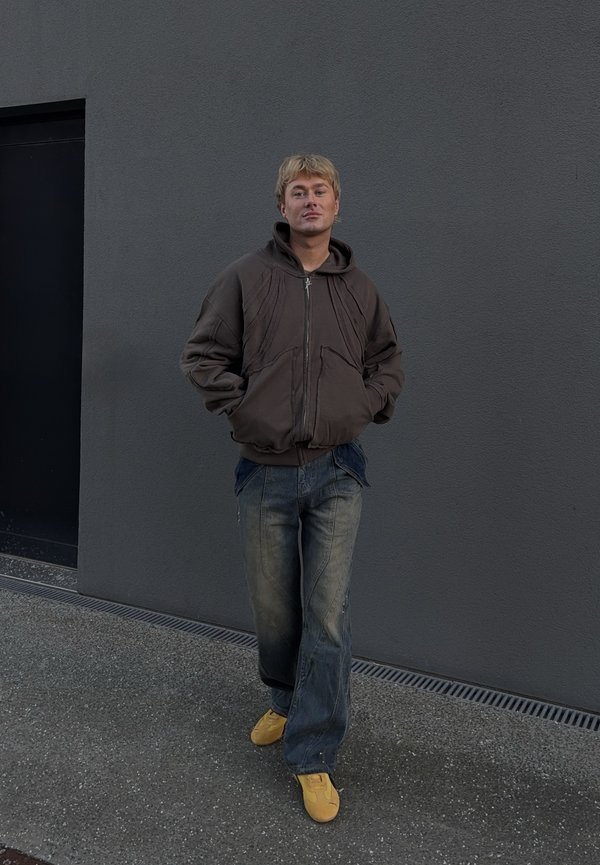 Ruda užtraukiama striukė su gaubtu, laisvos mėlynos džinsinės kelnės ir geltoni sportbačiai. Audinys minkštas, matomos siūlės ir laisvas prigludimas.