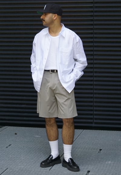 Camisa blanca de botón sobre una camiseta blanca de tirantes, combinada con pantalones cortos beige. Zapatillas negras, calcetines blancos y una gorra negra completan el atuendo.