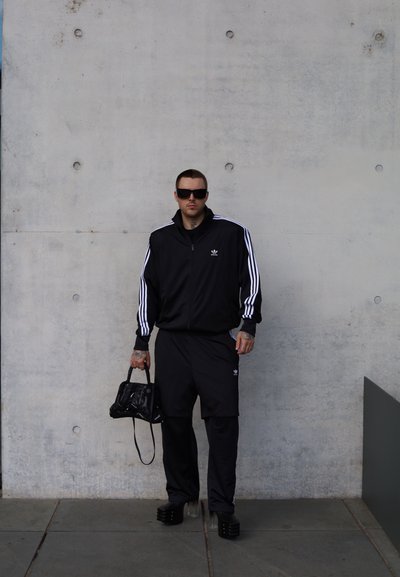 Chándal atlético negro con rayas blancas, camiseta de manga corta y pantalones cortos holgados. Bolso negro y calzado robusto. Fondo de concreto.