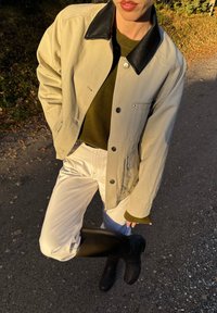 Beidzis žakets ar melnu ādas apkakli, zaļš džemperis, balti bikses un melnas ceļgala zābaki, viss fona ar gružu celi.