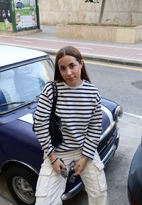 Suéter listrado em creme e azul-marinho, calças cargo brancas, segurando óculos de sol; de pé ao lado de um carro vintage azul; fundo de rua urbana.