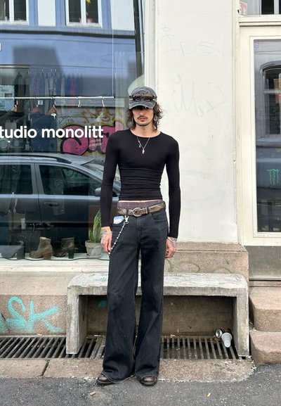 Camisa negra de manga larga ajustada, vaqueros negros de campana y una cadena con un llavero. Gafas de sol oscuras y una gorra gris. Se encuentra frente a un escaparate.