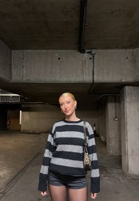 Černobíle pruhovaný svetr s dlouhými rukávy, nošený s černými šortkami. Modelka drží kabelku s leopardím potiskem. Beton pozadí.