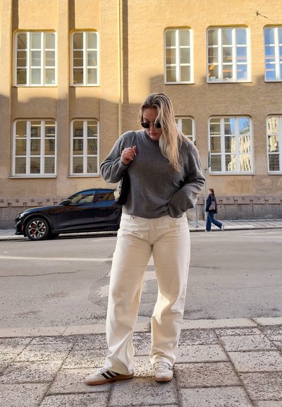 Suéter gris, pantalones de pierna ancha blancos y zapatillas con rayas negras. Bolso negro con correa de cadena. Fondo urbano con paredes amarillas y ventanas.