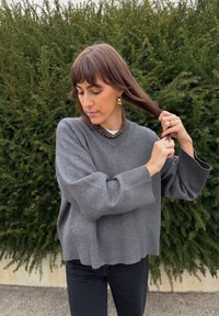 Maglione grigio oversize con scollo a V, polsini a coste, abbinato a una collana choker e orecchini a goccia. Sullo sfondo si vede della vegetazione.