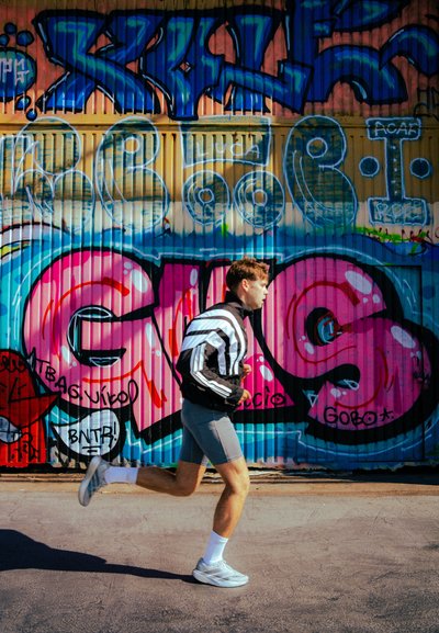 Corredor con una chaqueta a rayas en blanco y negro, pantalones cortos grises y zapatillas grises, moviéndose frente a una pared vibrante de graffiti con diseños en rosa y azul.
