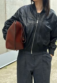 Svart läderbomberjacka med en ribbad krage, tillsammans med grå veckade byxor. Mörkbrun läderväska med slät textur.