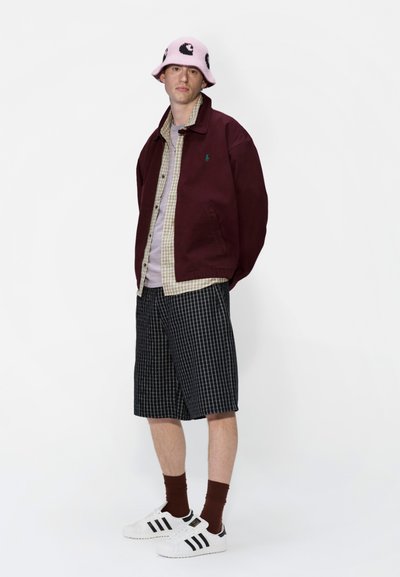 Chaqueta burdeos, camisa de cuadros con botones, sombrero tipo cubo rosa con estampado, pantalones cortos a cuadros, zapatillas blancas con rayas negras, calcetines marrones.