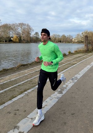Hombre con camisa verde de manga larga y pantalones negros trotando por un sendero junto al lago, con árboles y cielo nublado de fondo.