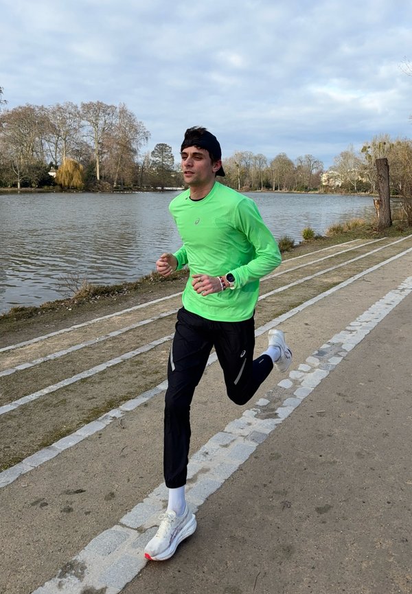 Homme en chemise verte à manches longues et pantalon noir en train de faire du jogging sur un sentier au bord du lac, avec des arbres et un ciel nuageux en arrière-plan.