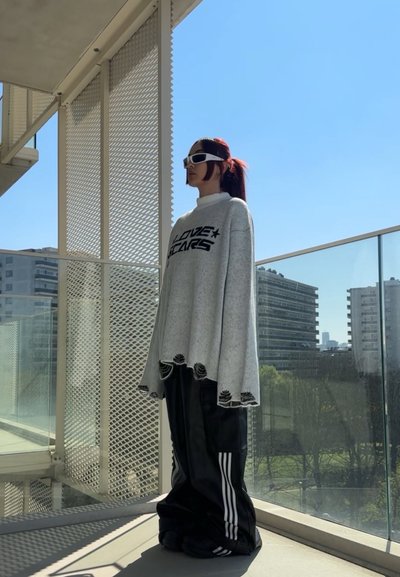 Suéter gris de gran tamaño con texto negro, dobladillo desgastado, combinado con pantalones de cuero sintético negro con rayas blancas y zapatos negros de gran tamaño.