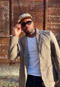 Homme portant un blazer à carreaux, une chemise blanche et une casquette, ajustant des lunettes de soleil foncées, debout devant un mur en bois vieilli sous la lumière du soleil.