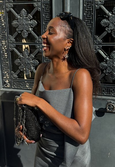 Mujer con cabello largo y gafas de sol en la cabeza ríe, sosteniendo un bolso negro, parada frente a una elaborada puerta de metal negra.