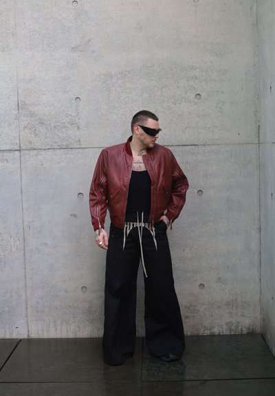 Hombre con gafas de sol negras, chaqueta de cuero roja, camiseta sin mangas negra y pantalones anchos negros, de pie contra una pared de hormigón.